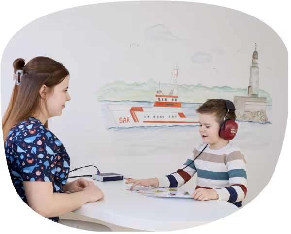 Ein Kind mit Kopfhörern sitzt an einem Tisch und spricht mit einer Frau, in einer Kinderarztpraxis.