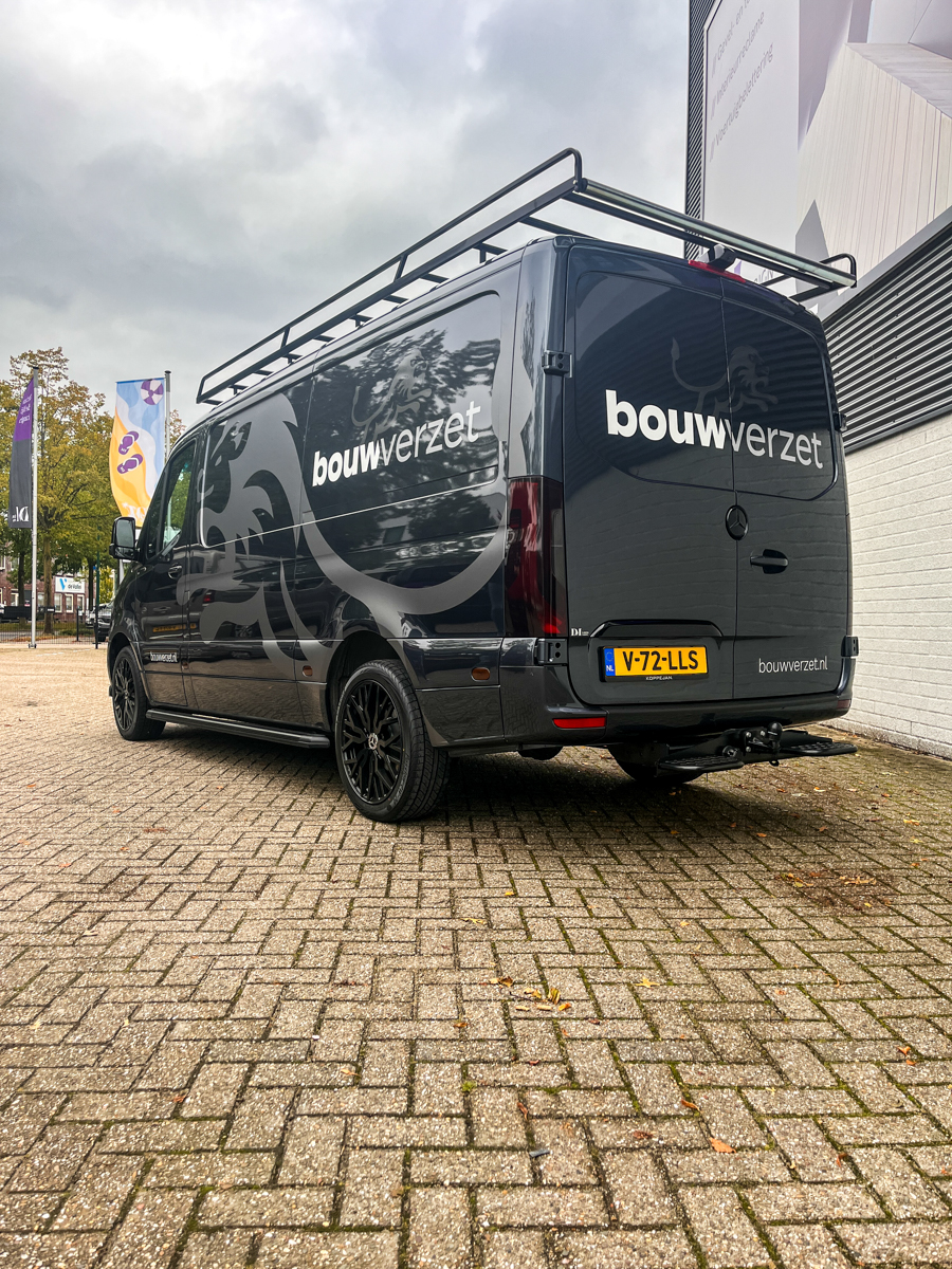 Zwarte bus met ‘bouwverzet’ bestickering geparkeerd op een bakstenen bestrating onder een bewolkte lucht.
