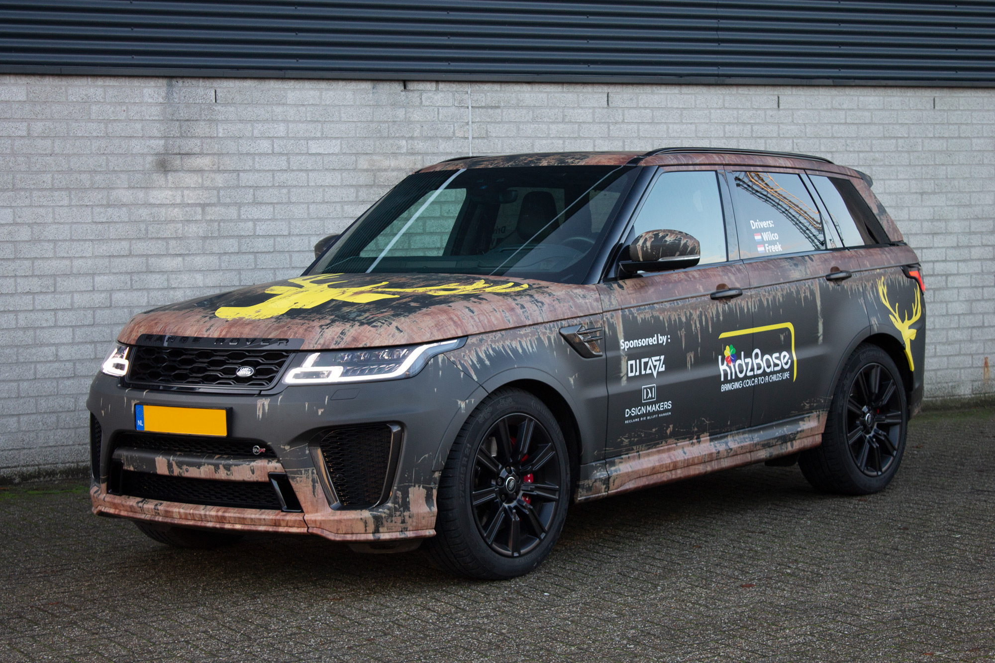 Een Range Rover SUV met een camouflagefolie in vuilmotief en sponsorlogo's staat geparkeerd tegen een grijze bakstenen muur.