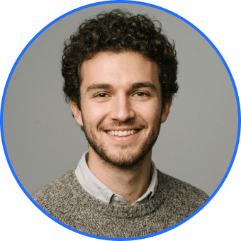 Smiling young man with curly hair and beard wearing a grey sweater and collared shirt on a grey background.
