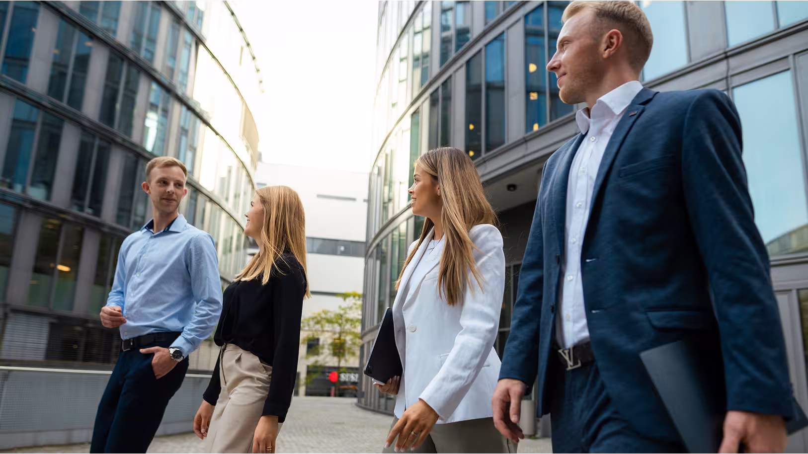 Vier junge Berufstätige in Business-Kleidung laufen gemeinsam durch einen modernen Bürokomplex mit Glasfassaden und unterhalten sich dabei.