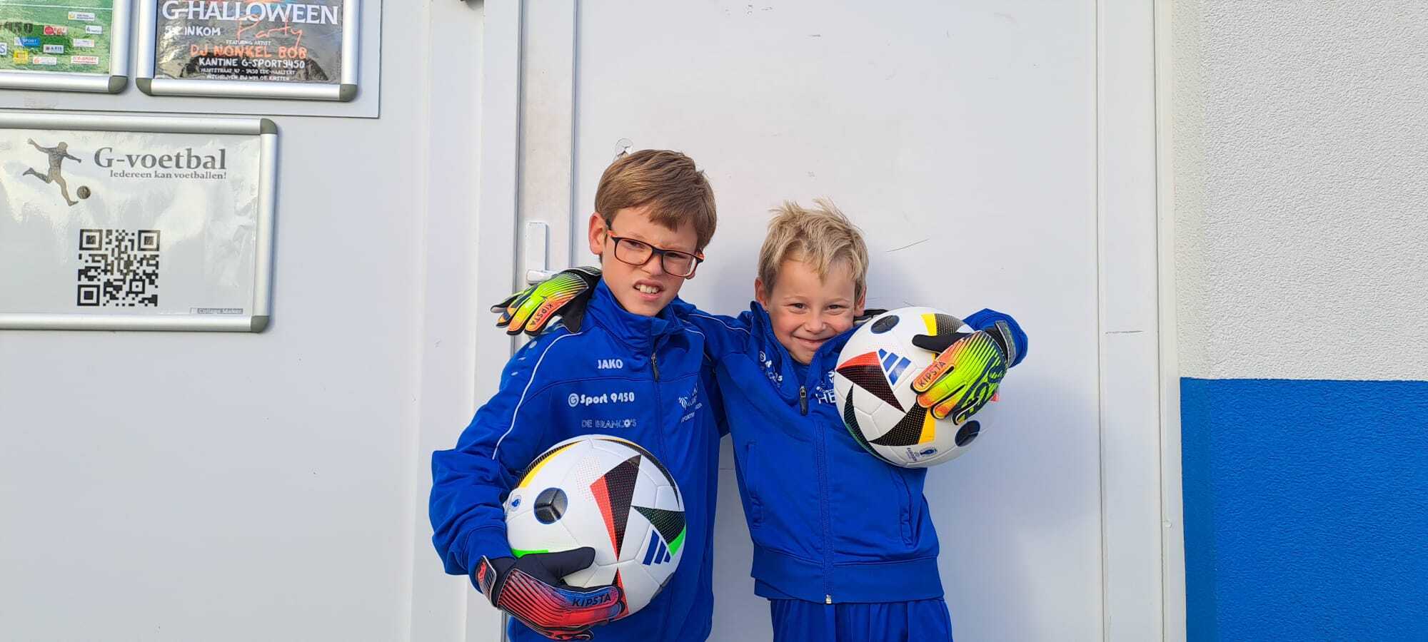Twee jonge kinderen met voetbalkleren en een voetbal in de hand poseren met een lach, war wijst op een sportieve en vrolijke omgeving