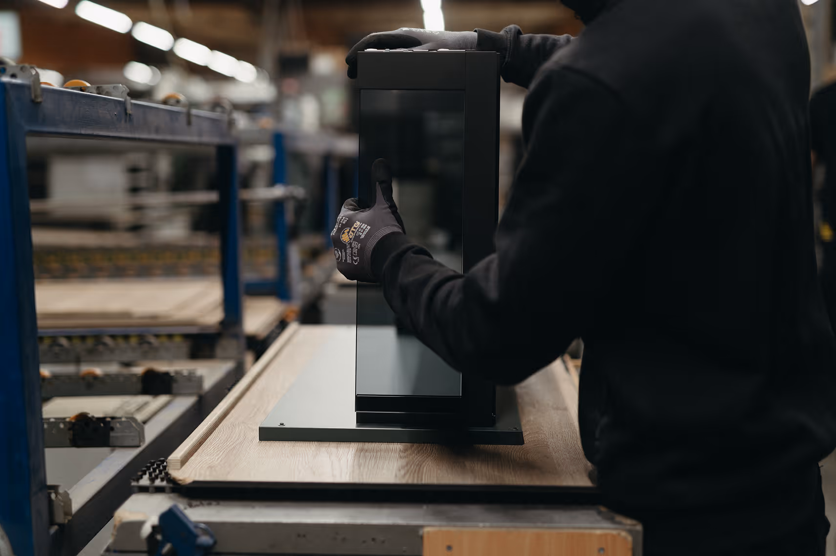 Person wearing black gloves assembling or inspecting a black metal frame on a factory conveyor belt.