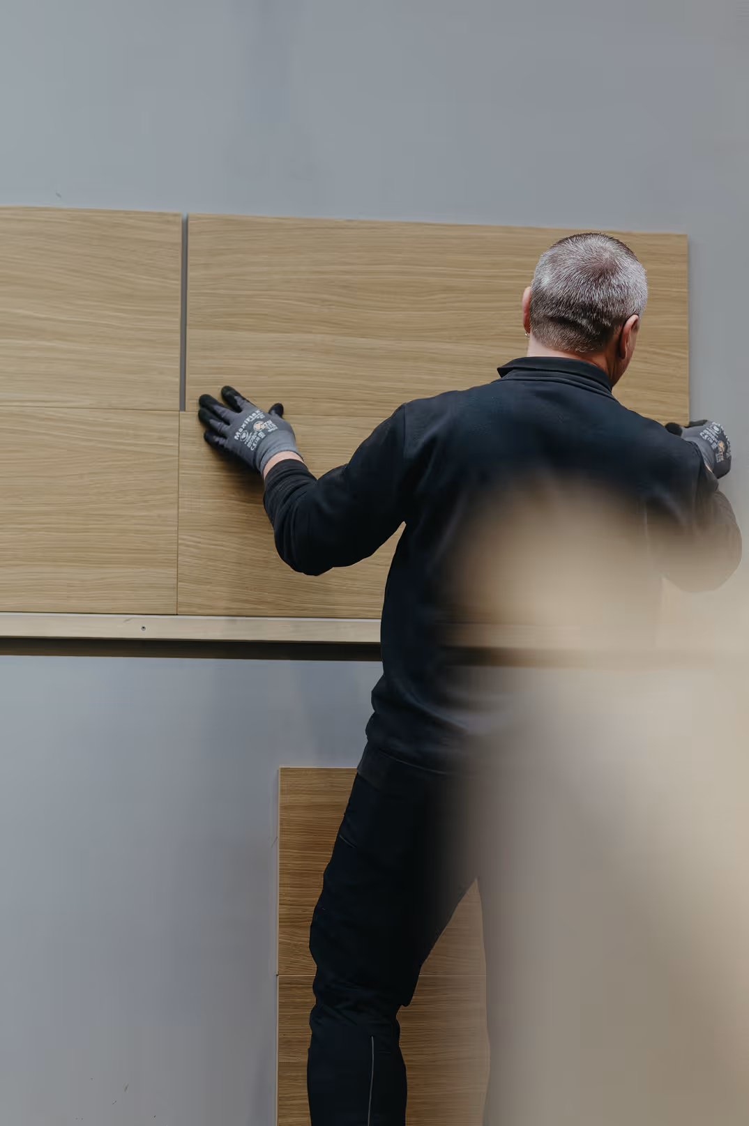 Ein Mann mit Arbeitshandschuhen montiert Holzwandverkleidung auf grauer Wand.