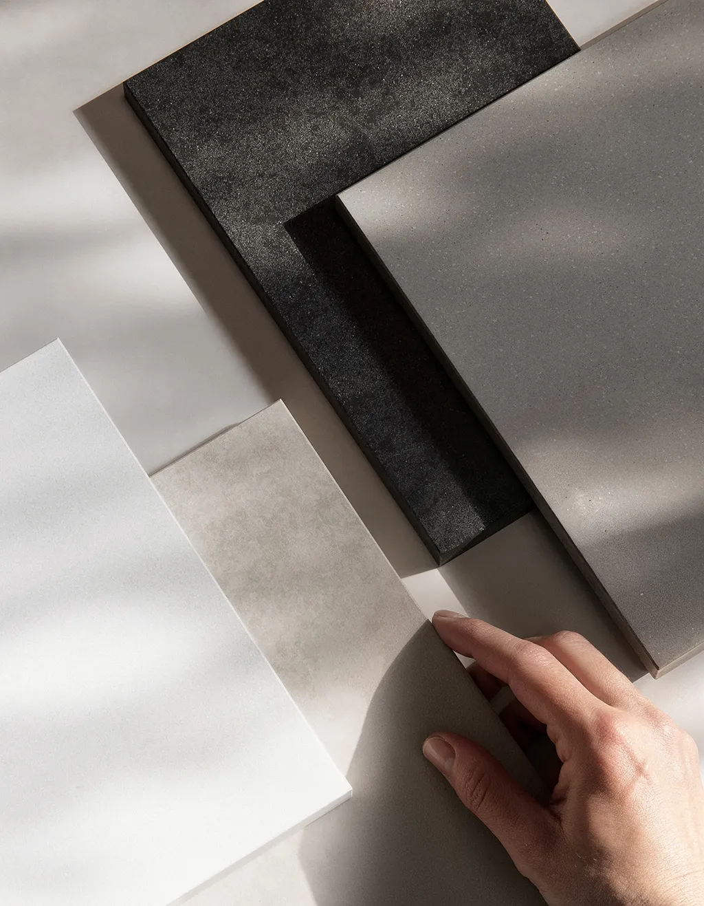 Hand berührt Musterkarten aus Steinplatten in Weiß, Beige, Grau und Schwarz auf weißem Hintergrund.