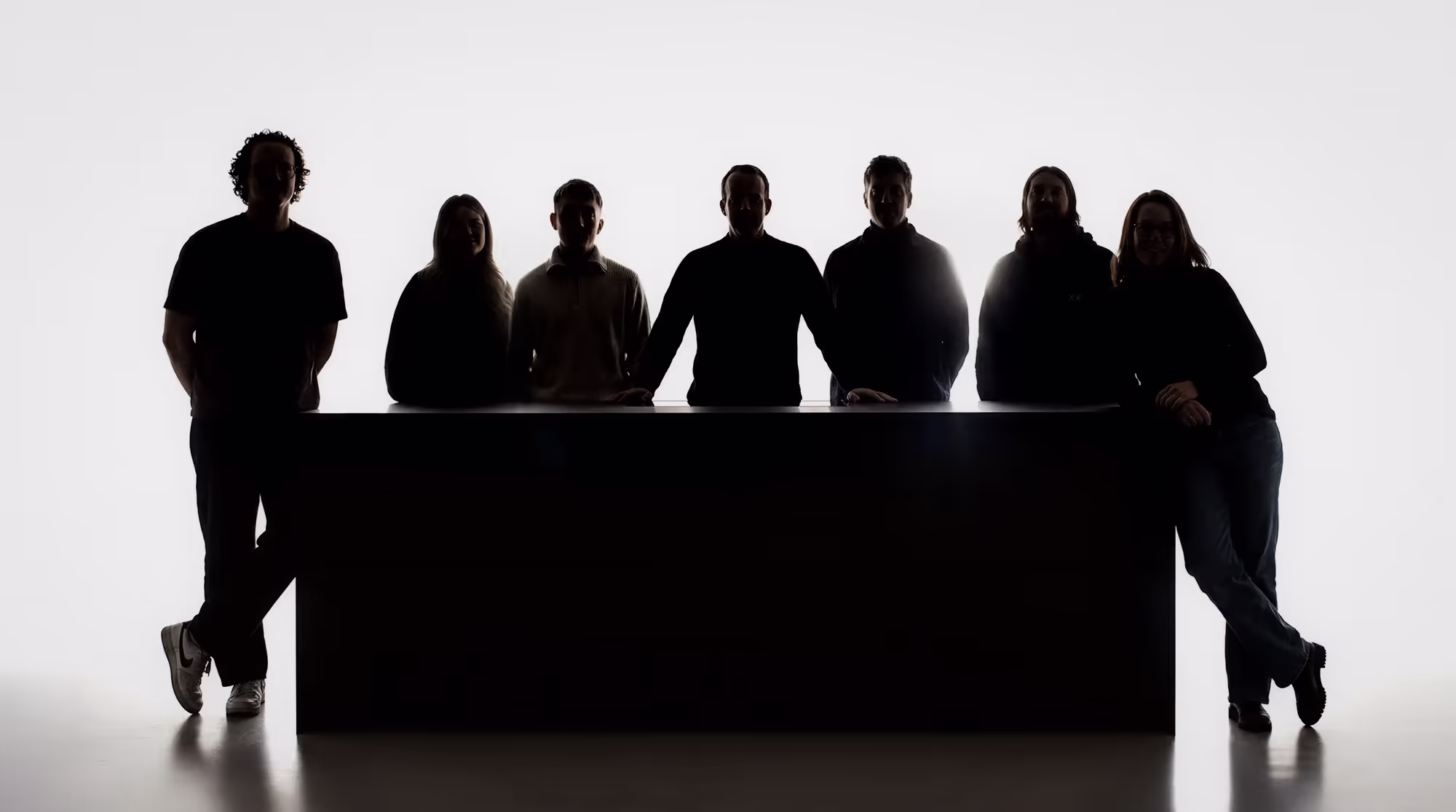 Schattenriss von sieben Personen, die hinter einem großen schwarzen Tisch stehen oder lehnen.