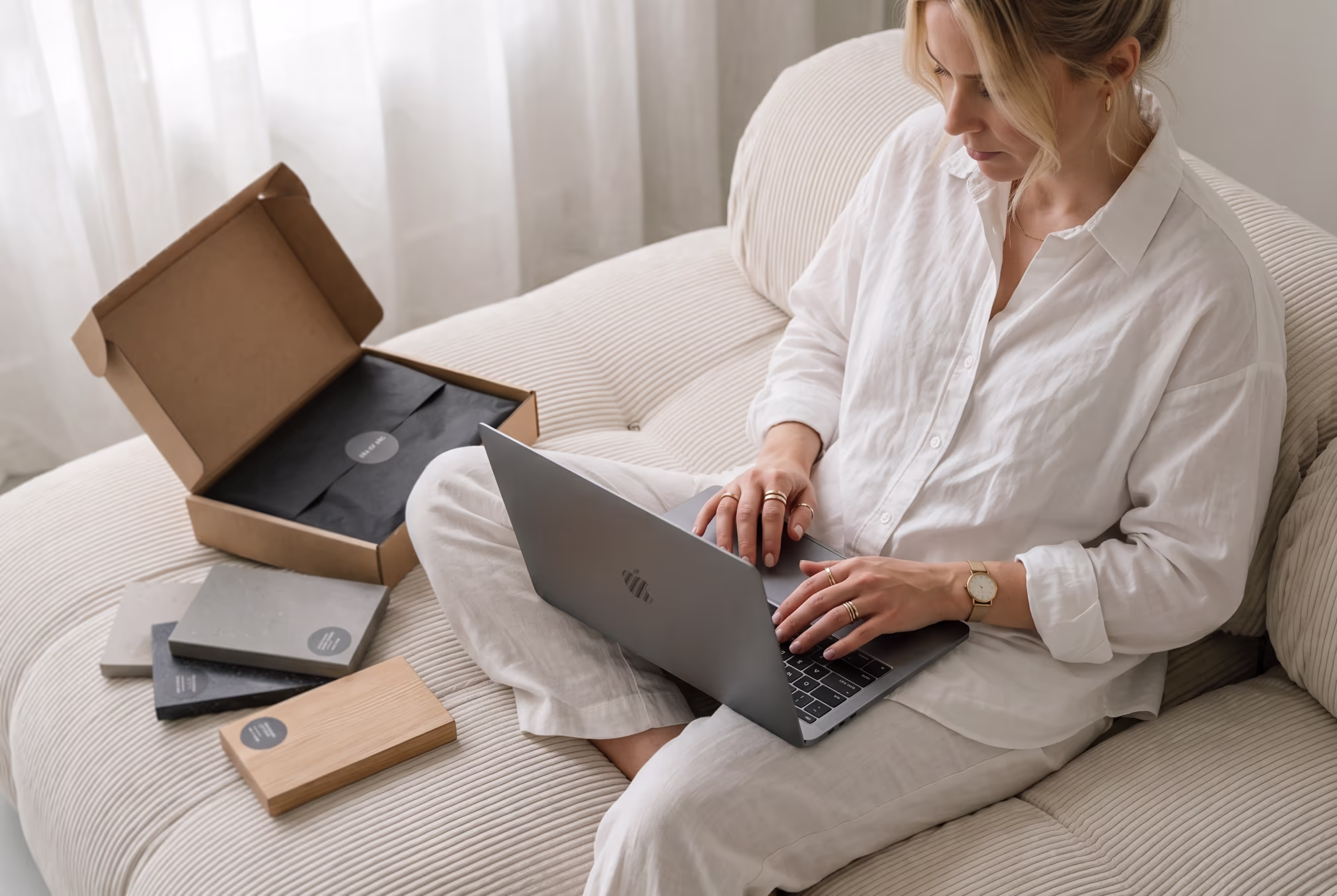 Frau in weißem Outfit arbeitet auf Laptop sitzend auf beigem Sofa, Musterproben liegen daneben.