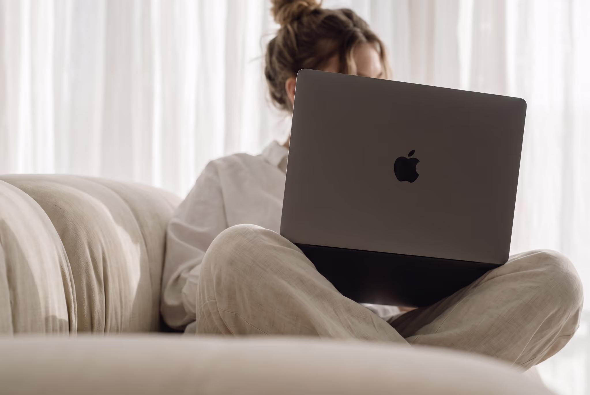 Person sitzt mit verschränkten Beinen auf einem beigen Sofa und benutzt einen Apple Laptop.