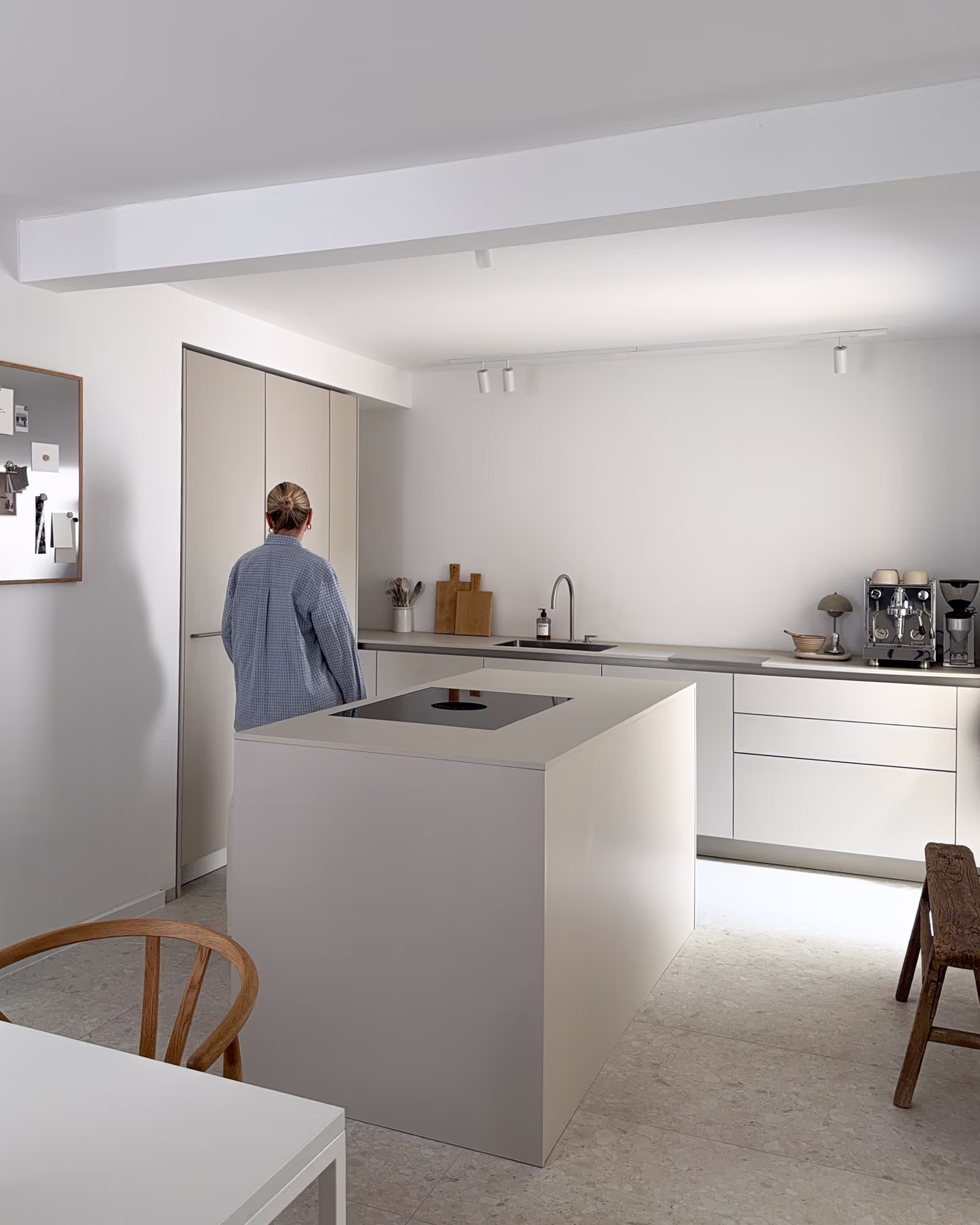 Eine moderne, minimalistische Küche mit einer zentralen Kücheninsel, eingebautem Kochfeld, weißen Schränken und einer Person mit grauem Mantel, die mit dem Rücken zur Kamera vor einem Schrank steht.
