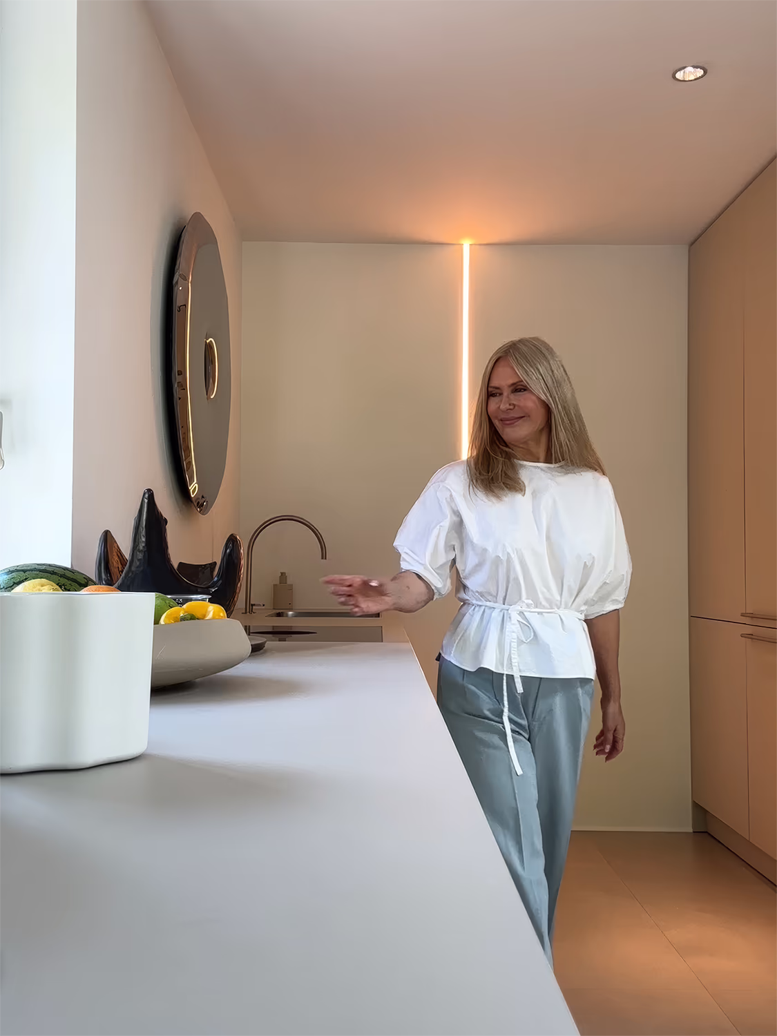 Frau mit weißen Kurzarmbluse und grauer Hose lehnt entspannt an einer modernen Küchenarbeitsplatte mit Obstschale, rundem Spiegel an der Wand und minimalistischem Design.