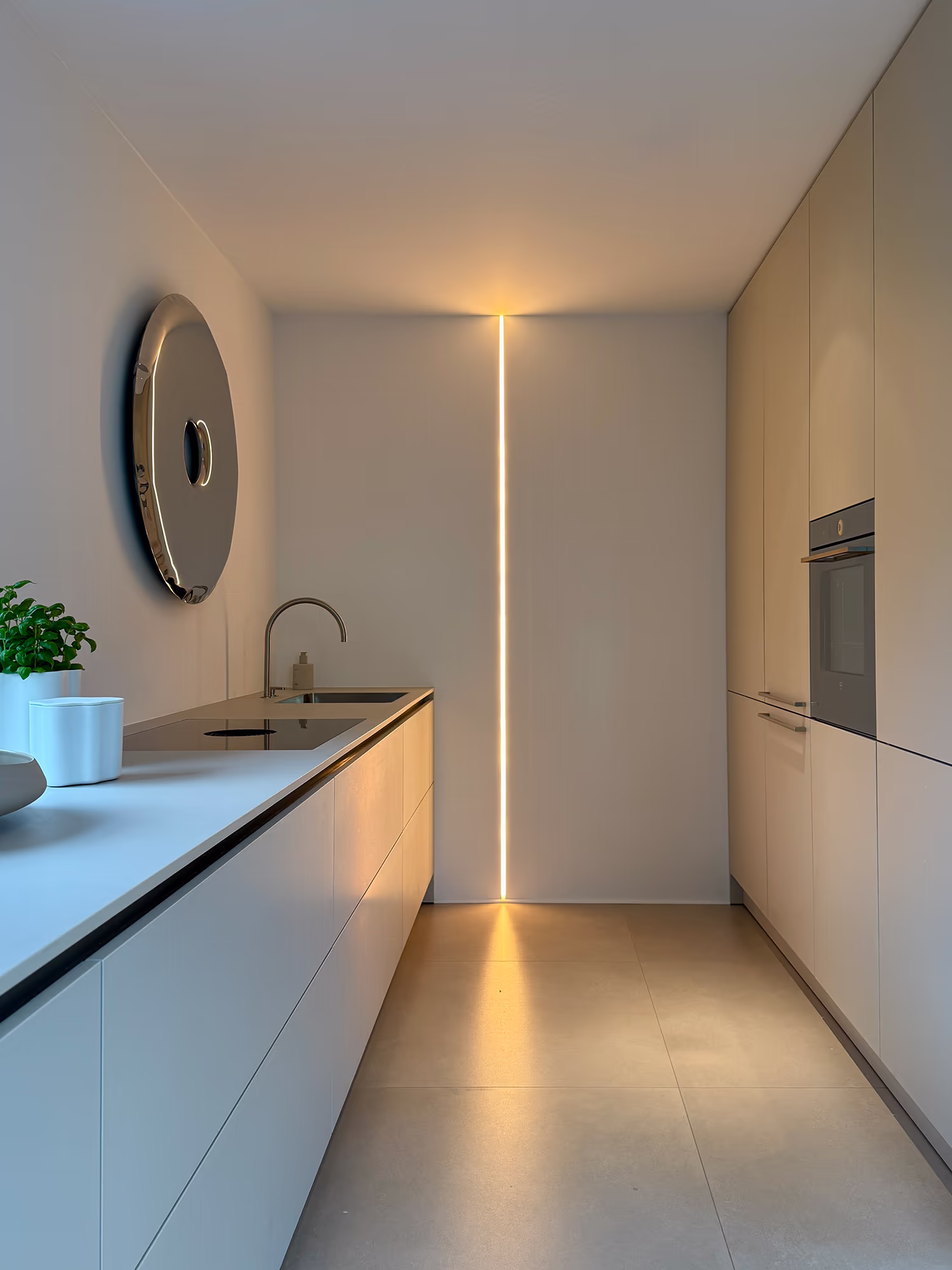 Moderne minimalistische Küchenzeile mit weißen Schränken, integriertem Backofen rechts, Spüle links, einer Pflanze und einer runden Wanddekoration, beleuchtet durch einen schmalen, vertikalen Lichtstreifen an der Wand.