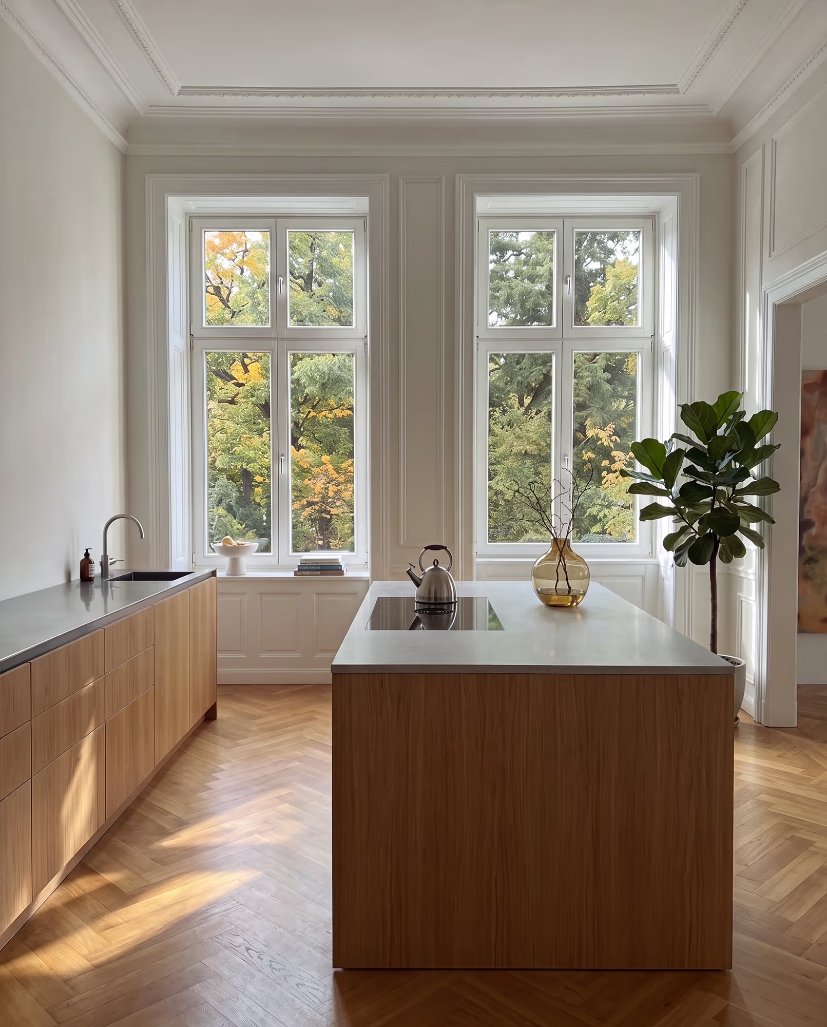 Helle, moderne Küche mit zwei großen Fenstern, weißen Wänden und Holzböden im Fischgrätmuster, einer Kücheninsel mit Kochfeld und Teekessel, Holzschränken, Spüle und grüner Pflanze in der Ecke.