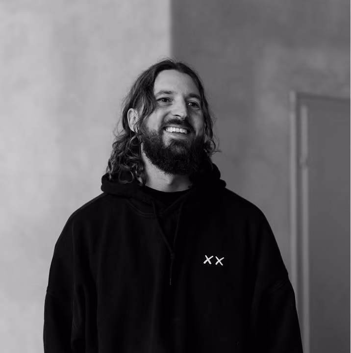 Schwarz-weiß-Foto eines lächelnden Mannes mit langem, lockigem Haar und Bart, der einen Kapuzenpullover trägt.