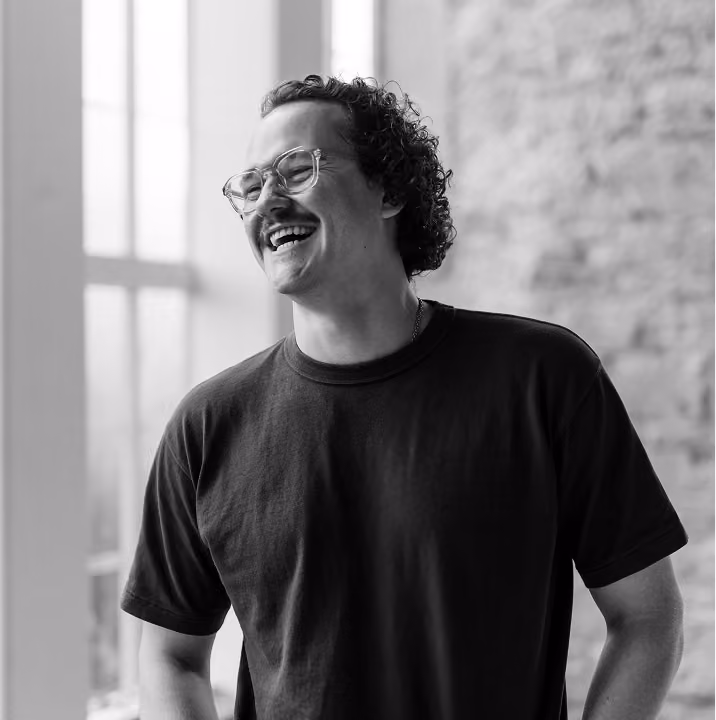 Schwarz-weiß-Foto eines lächelnden Mannes mit lockigem Haar, Schnurrbart und Brille, der ein schlichtes dunkles T-Shirt trägt, mit verschwommenem Fenster und Backsteinmauer im Hintergrund.