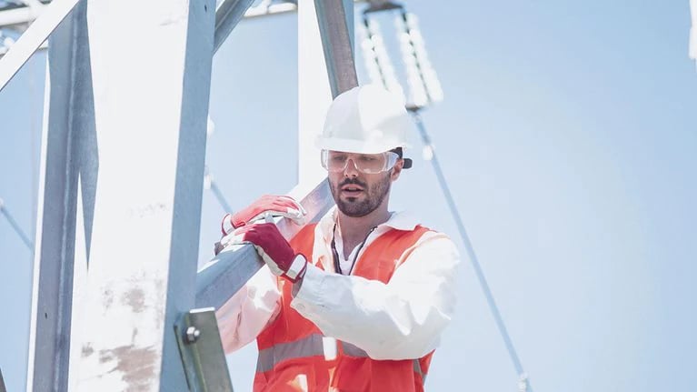 Construction worker wearing a white hard hat, safety glasses, and an orange safety vest working on a metal structure.