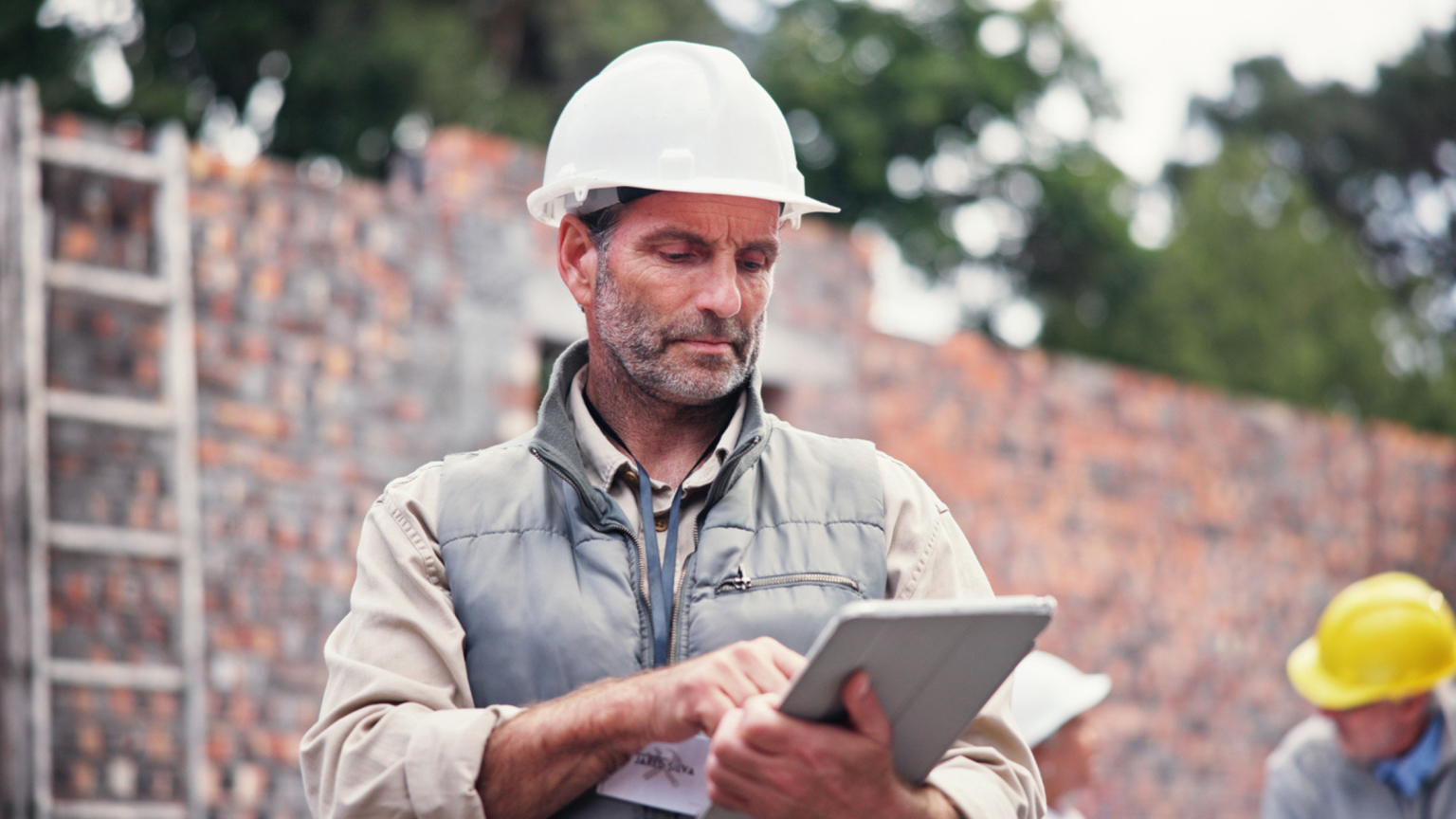 Construction worker wearing a white hard hat and gray vest using a tablet at a brick wall construction site.