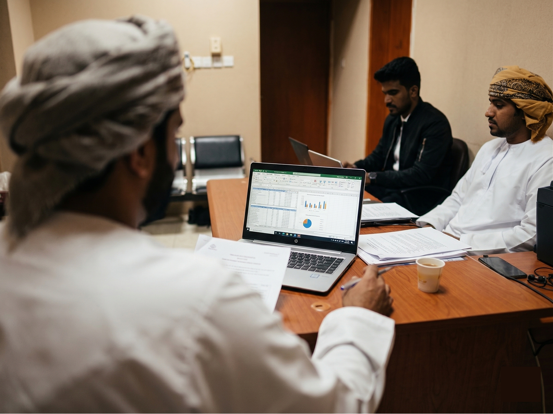 Three men in traditional Middle Eastern attire in a meeting room working on laptops and reviewing documents at a wooden table.