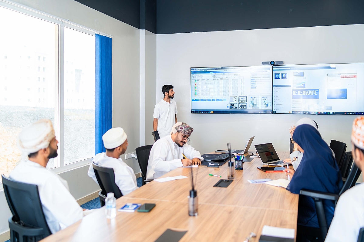 Group of people in traditional attire attending a meeting in a conference room with two large screens displaying digital data.
