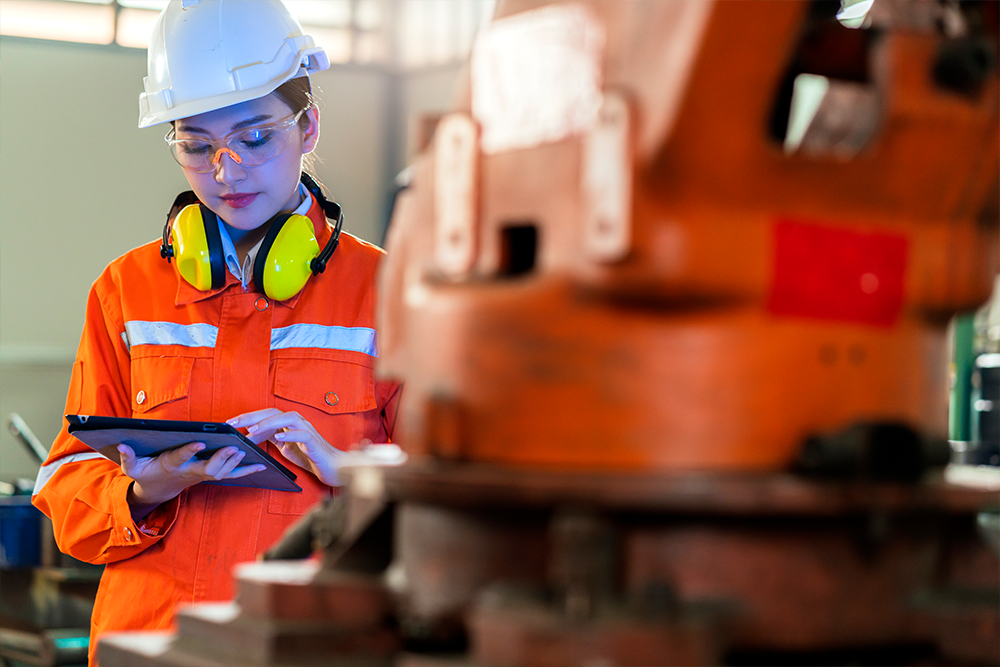 Female engineer in orange safety jacket, white hard hat, and yellow ear protectors using a tablet near industrial machinery.