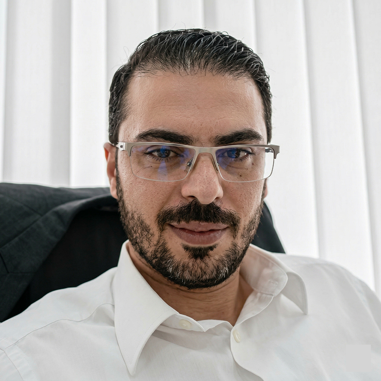 Man with dark hair, beard, and glasses wearing a white shirt sitting in front of white curtains.