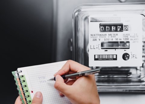 Person writing notes with a pen in a notebook while reading an analog electricity meter.