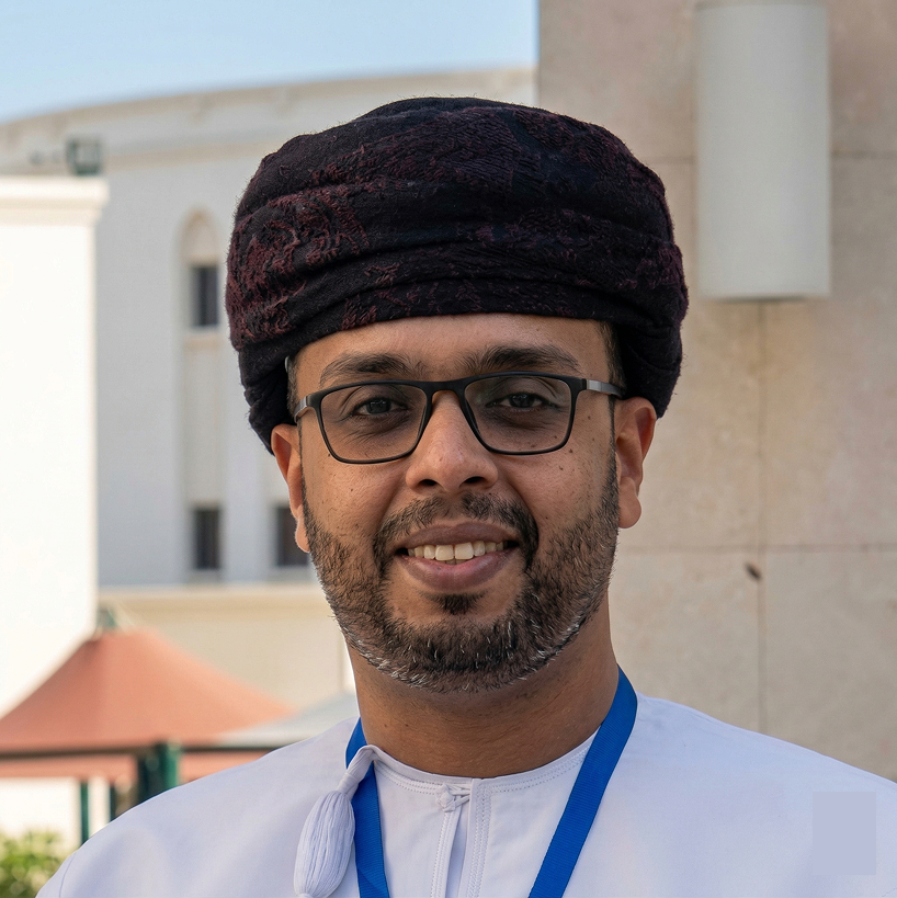 Smiling man wearing glasses and a traditional dark turban with a white garment and blue lanyard outdoors.