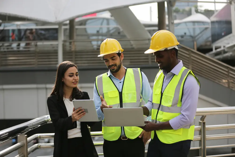 Three professionals, two men in yellow safety helmets and vests and a woman in business attire, discussing while holding a laptop and a tablet outdoors.