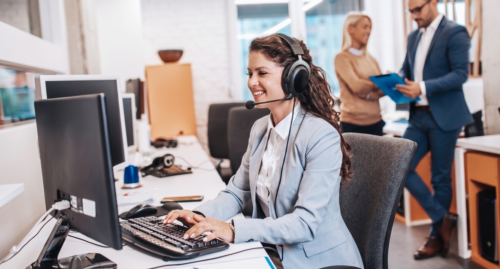 Smiling woman wearing a headset and typing on a keyboard in a modern office with two colleagues discussing in the background.