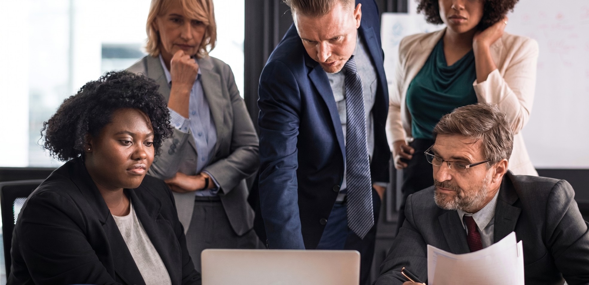 Diverse group of five business professionals intensely discussing work around a laptop in a modern office.