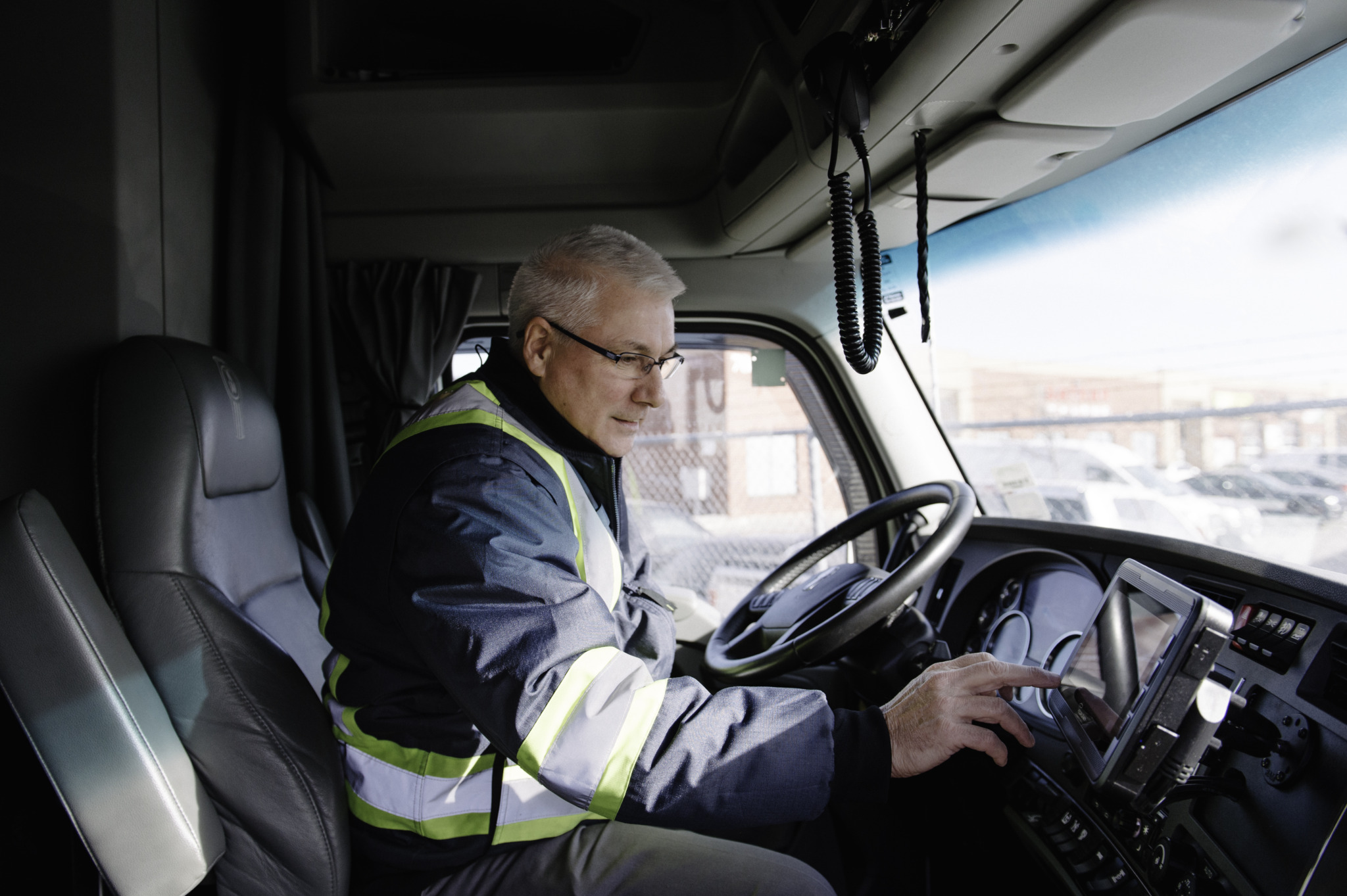Older man wearing glasses and a reflective safety jacket operating a touchscreen device inside a truck cab.