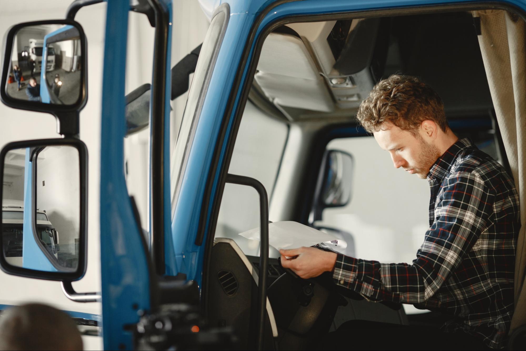 Man in a plaid shirt sitting inside a blue truck, looking intently at a paper checklist.