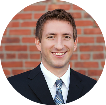 Smiling man in a black suit, white shirt, and striped tie against a red brick wall.