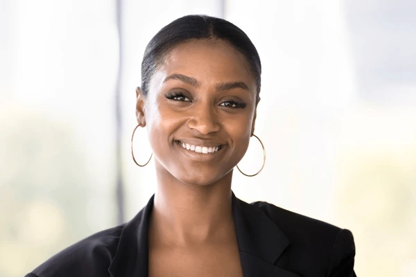 Smiling woman with hoop earrings and dark blazer in softly lit indoor setting.
