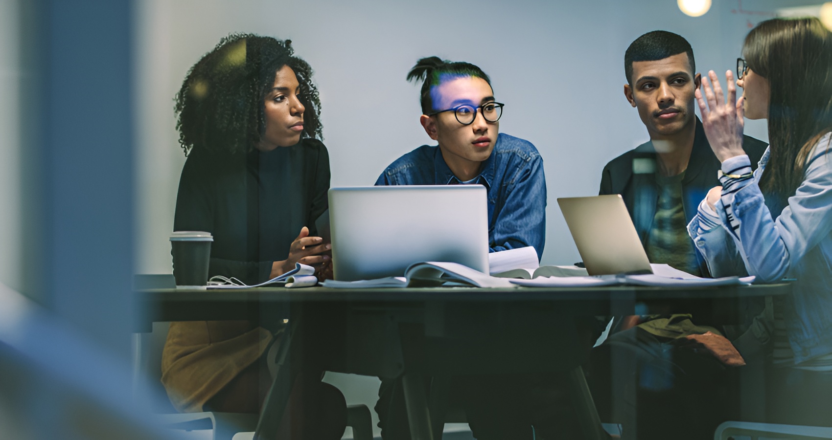 Four diverse young professionals engaged in a discussion around a table with laptops and open books.