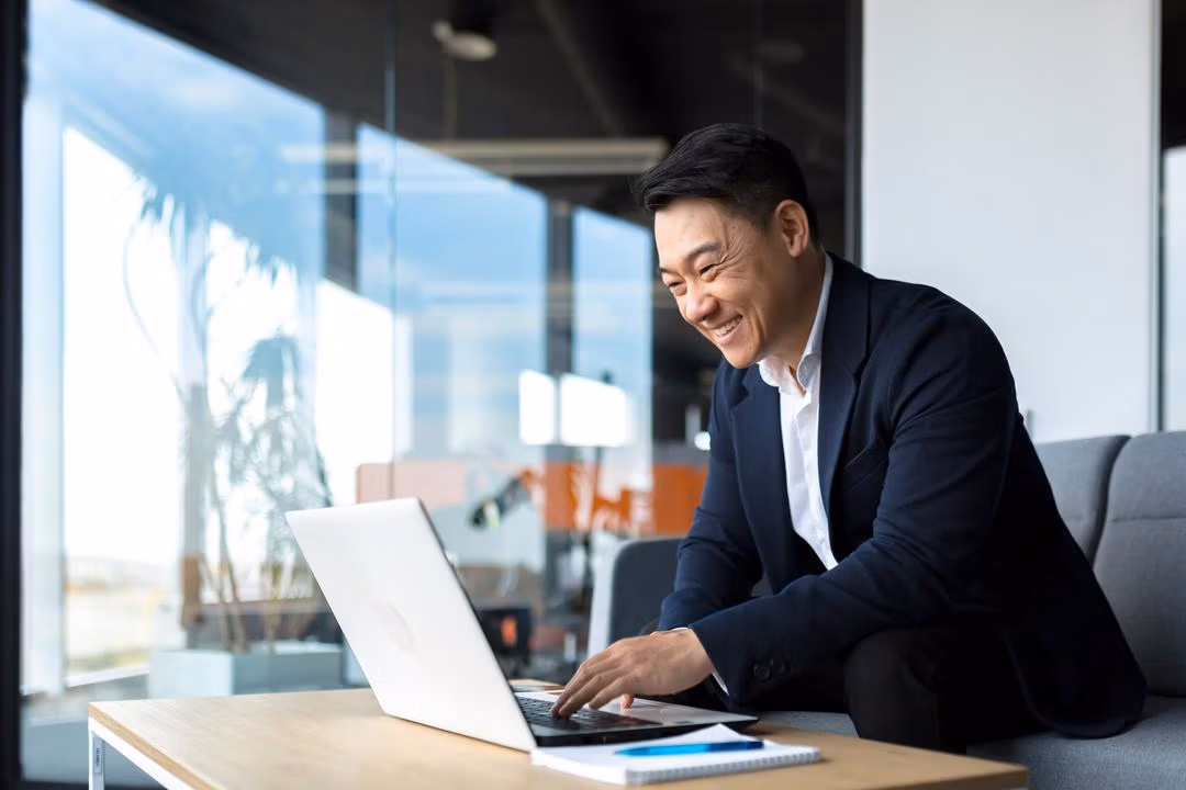 Smiling man in business attire working on a laptop at a wooden table in a modern office with large windows.