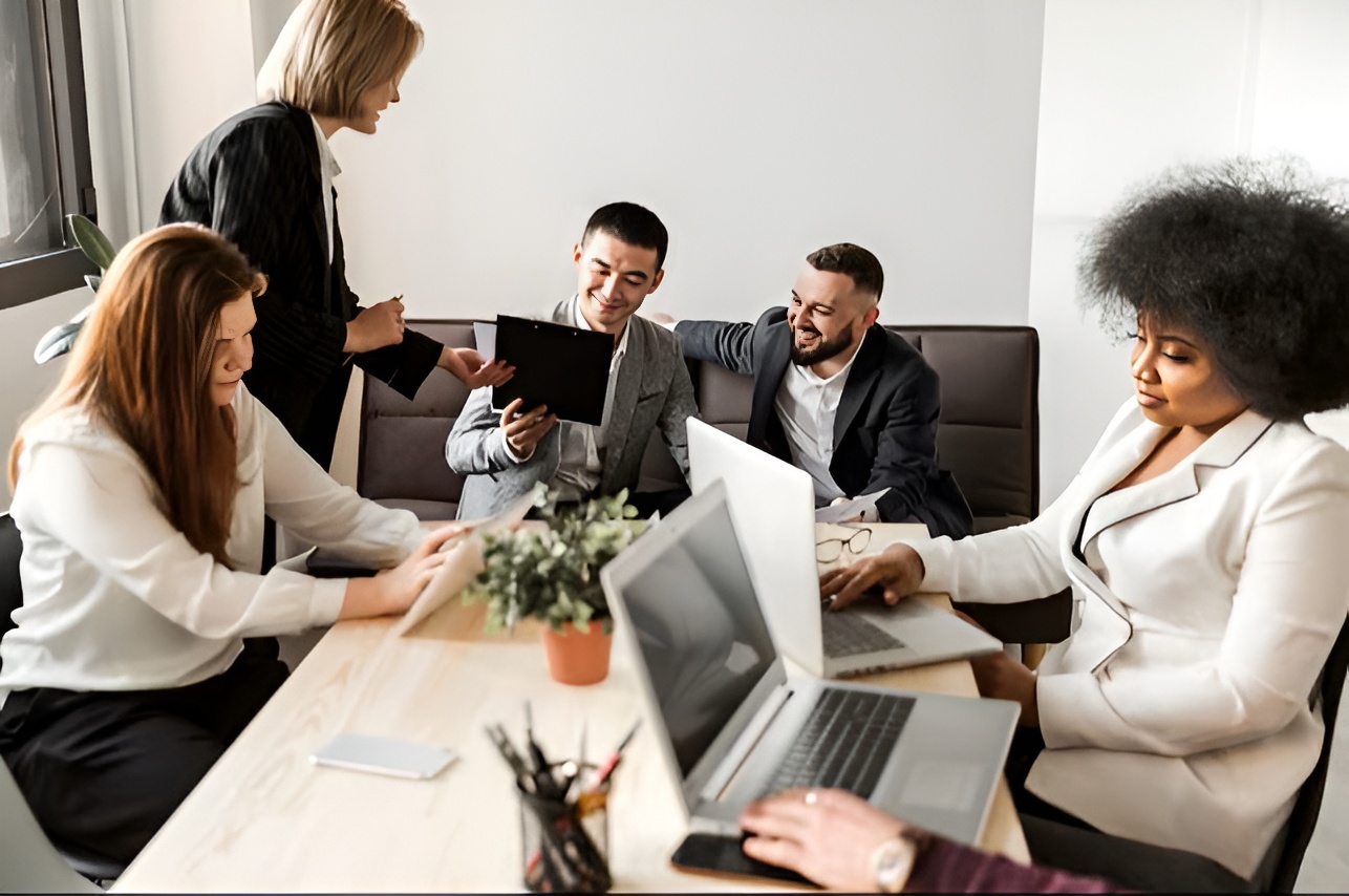Five diverse colleagues in business attire having a meeting around a table with laptops and documents.