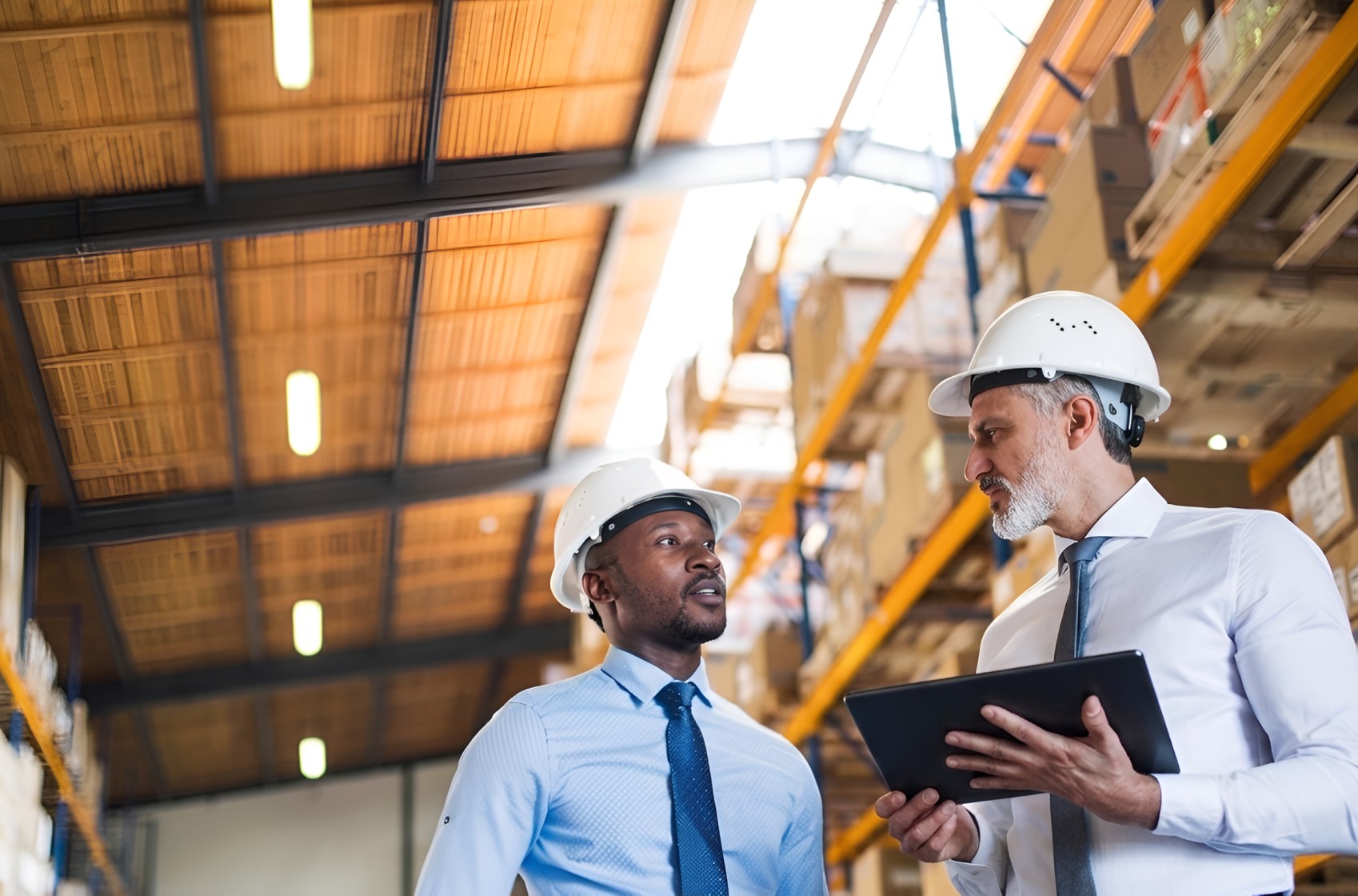 Two men in hard hats discussing work inside a warehouse with shelves stocked with boxes, one holding a tablet.