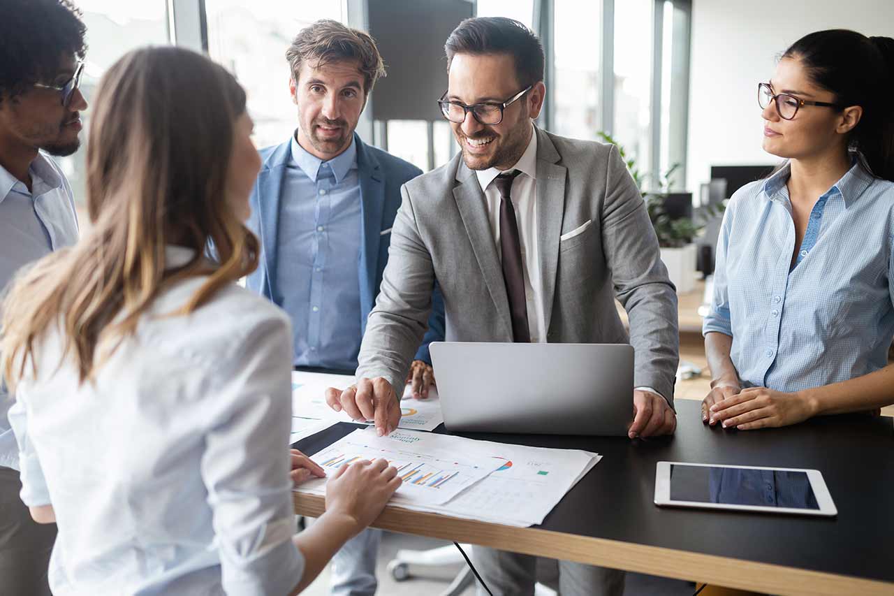 A diverse group of five professionals in an office discussing charts and data on a table with a laptop and tablet.