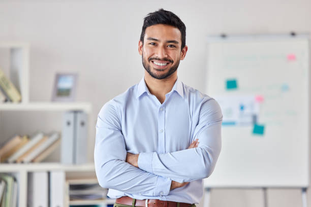 Smiling man in a light blue shirt with arms crossed standing in an office setting with a blurred whiteboard in the background.