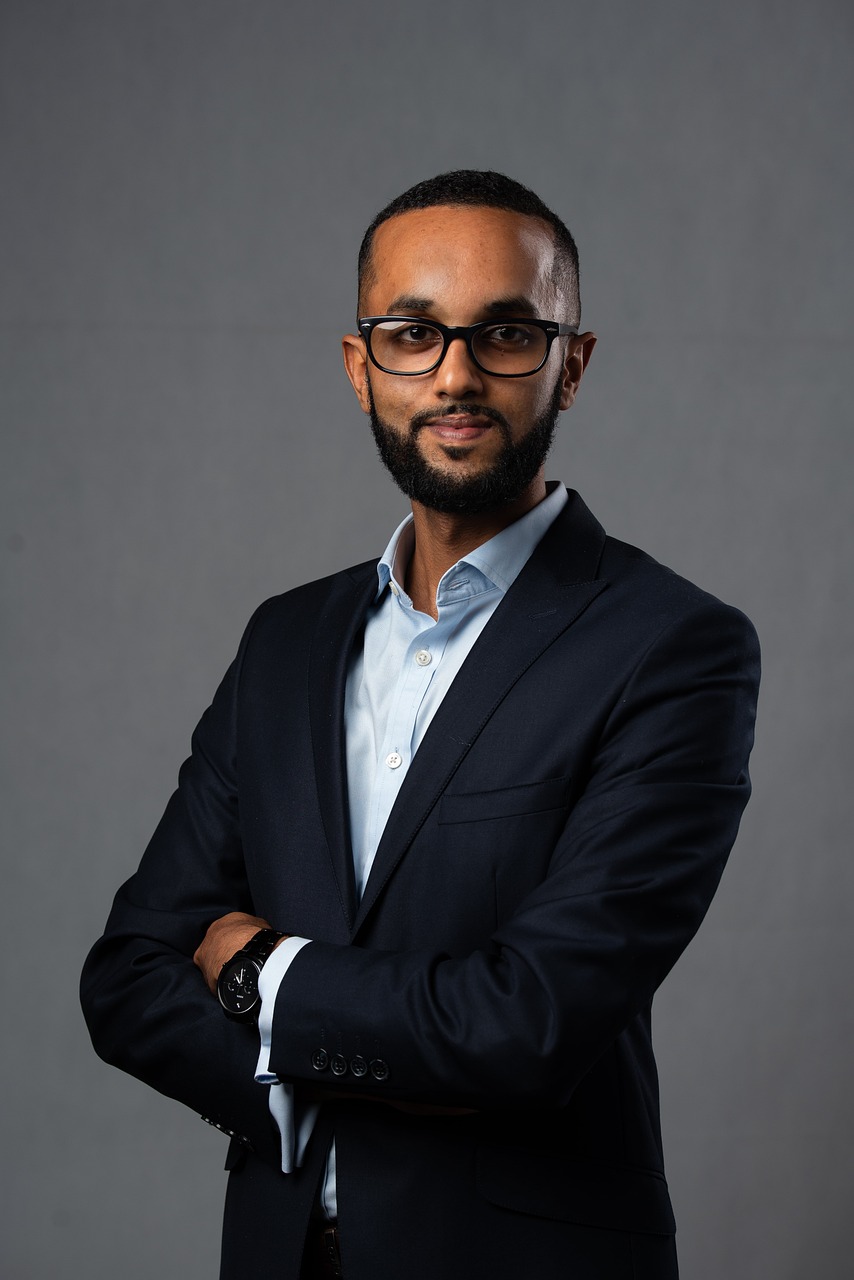 Man with beard and glasses wearing a dark suit and light blue shirt, standing with arms crossed against a gray background.