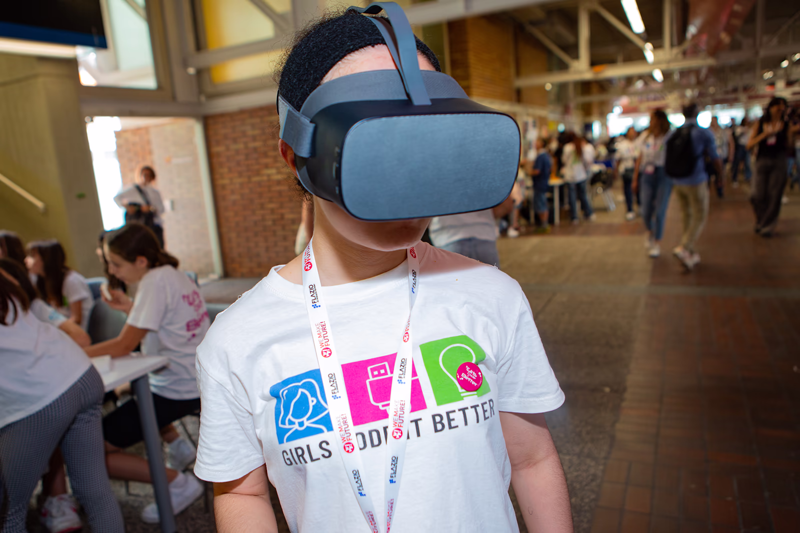 Ragazza indossa un visore VR con maglietta "Girls Code It Better" in un ambiente affollato.