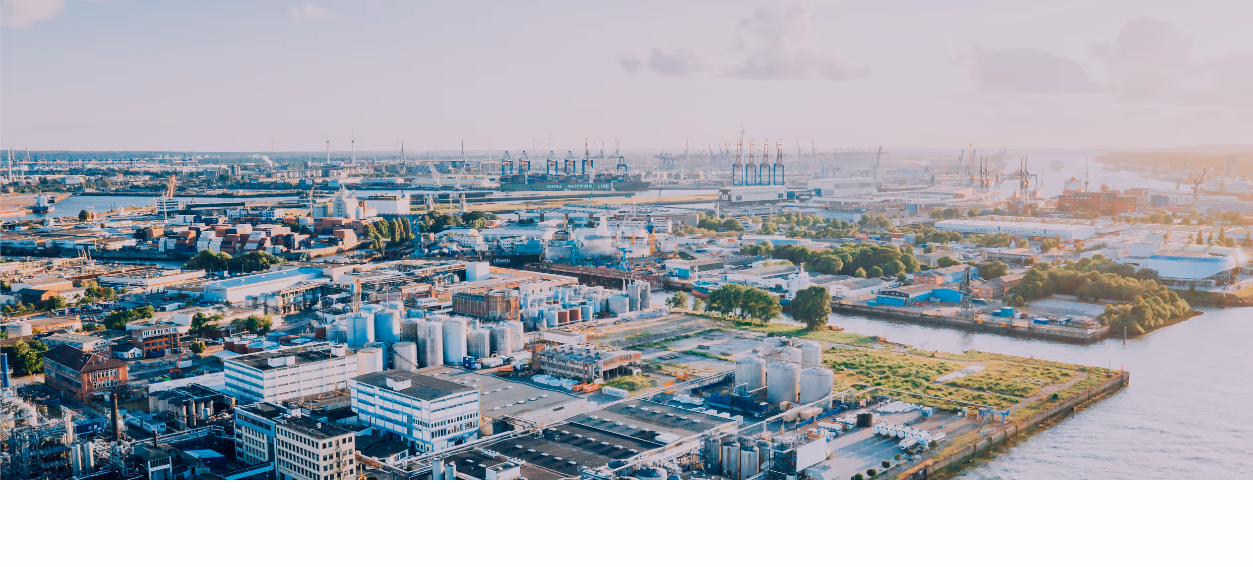 Aerial view of a large industrial port with warehouses, storage tanks, shipping containers, cranes, and water channels under a cloudy sky.
