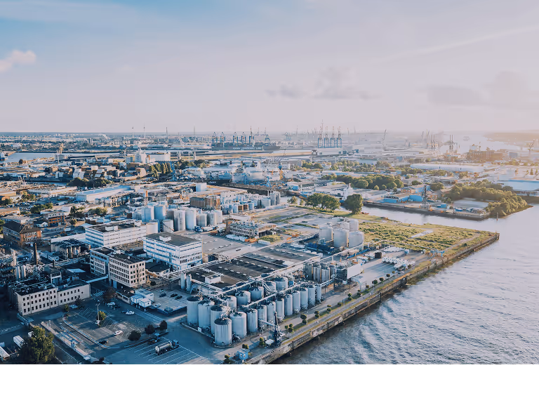 Aerial view of a large industrial port with warehouses, storage tanks, shipping containers, cranes, and water channels under a cloudy sky.