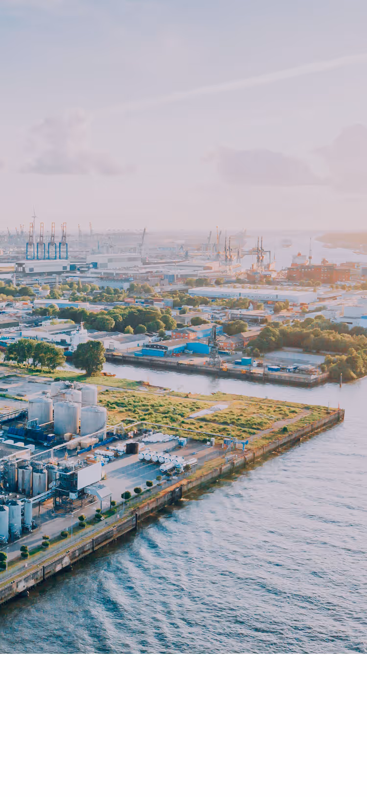 Aerial view of a large industrial port with warehouses, storage tanks, shipping containers, cranes, and water channels under a cloudy sky.