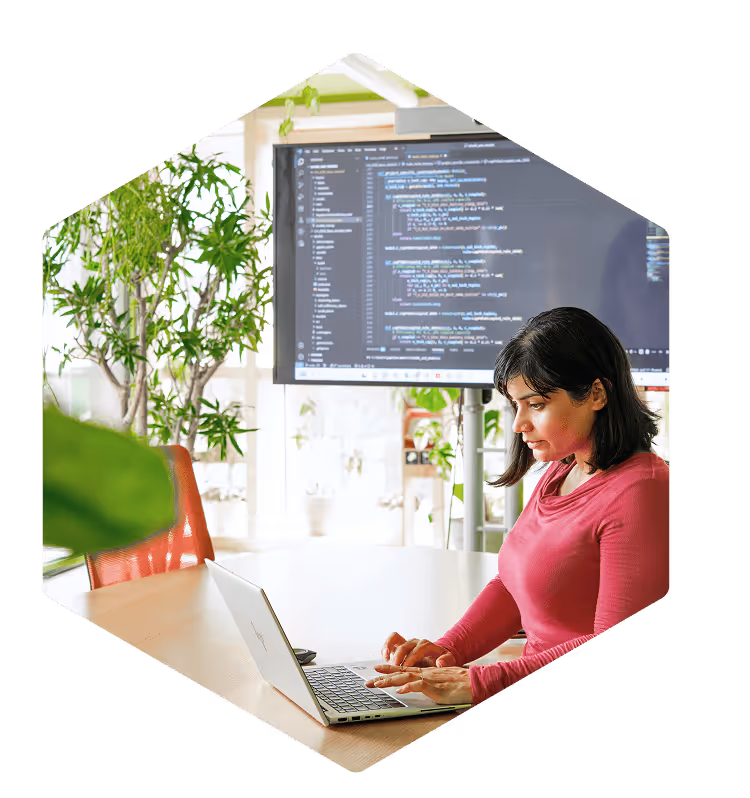 Woman in a pink long-sleeve top working on a laptop at a table with a large screen behind her displaying programming code.