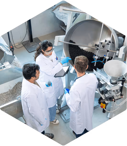 Three scientists in white lab coats inspecting black CO2 pellets in a large industrial machine of the EMPA in a laboratory setting.