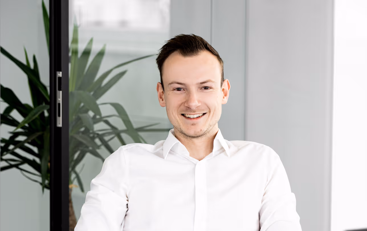 Junger Mann mit weißen Hemd lächelt in einem modernen Büro mit Pflanzen im Hintergrund.