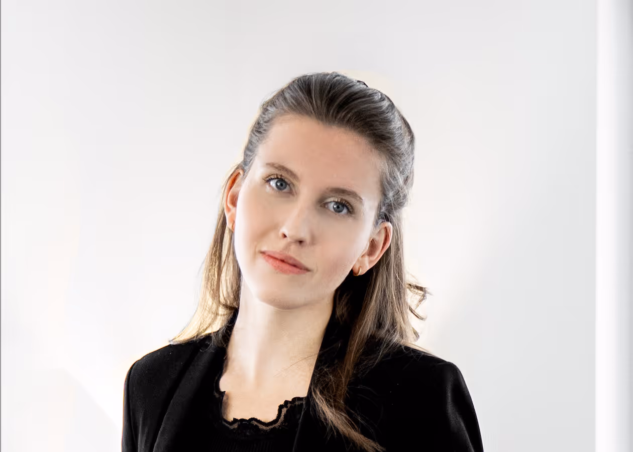 Porträt einer Frau mit blauen Augen und braunen Haaren, die eine schwarze Bluse und einen schwarzen Blazer trägt, vor hellem Hintergrund.