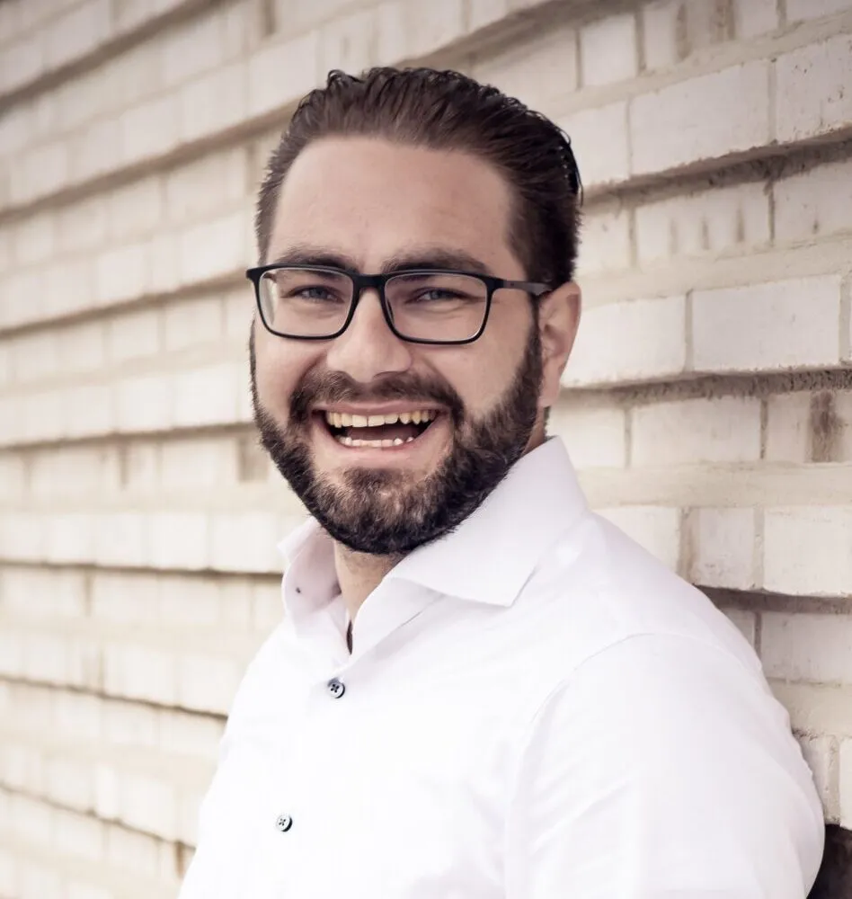 Ein lachender Mann mit Brille, dunklem Haar und Bart in einem weißen Hemd vor einer weißen Backsteinwand.