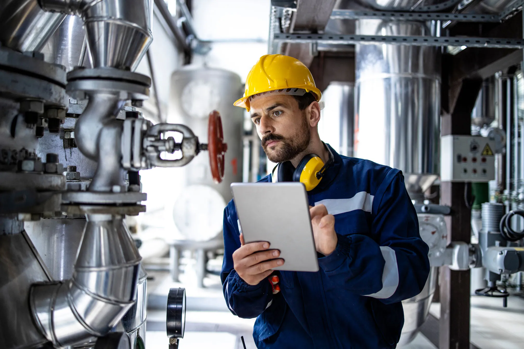 Techniker mit gelbem Helm und Tablet überprüft industrielle Rohrleitungen und Ventile in einer Fabrik.
